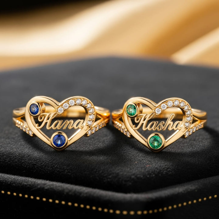 Two gold heart-shaped rings with gemstones and names on a black surface.